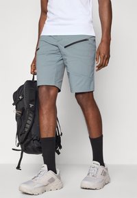 Shorts gris clair avec une texture lisse, dotés de deux poches zippées et de coutures structurées. Associés à des baskets blanches et des chaussettes noires.