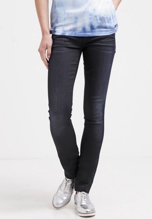 Pantalones ajustados de mezclilla azul oscuro con una textura suave y un brillo sutil, combinados con unos oxford plateados con detalles perforados.