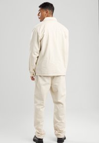 Beige katoenen jas met een rechte snit, knoopmanchetten en zijsplitten, gecombineerd met bijpassende taps toelopende broek en zwarte schoenen.