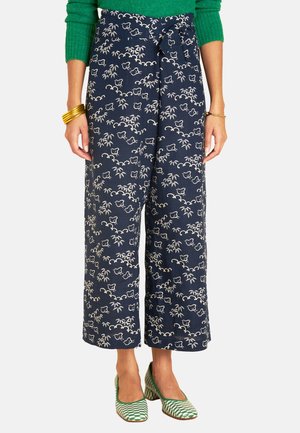 Weite Hosen in Marineblau mit einem weißen Blumen- und Bambusmuster. Leichter Stoff mit einem gebundenen Bund. Das Model trägt grüne Schuhe mit geometrischem Design.