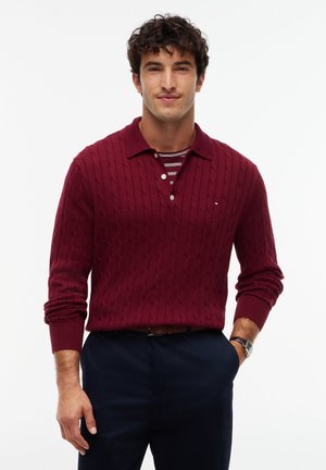 Mand iført en burgundy kabelstrikket sweater over en stribet skjorte, med mørke bukser og et armbåndsur, står mod en ensartet hvid baggrund.