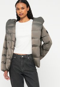 Doudoune en nylon gris atténué avec une finition brillante, dotée d'une capuche et de fermetures à pression. Portée par-dessus une chemise blanche et un jean noir.