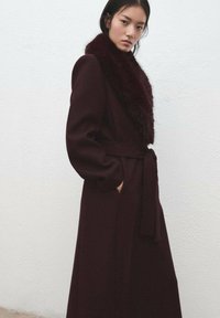 Bordeauxrode lange jas met een getextureerde nepbontkraag, voorzien van een ceintuur en zijzakken, gladde stof, getailleerde pasvorm.