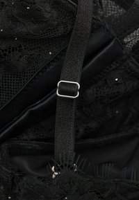Detail van een zwart kanten kledingstuk met een satijnen band met een zilveren metalen slider, een gestructureerde kanten overlay en kleine decoratieve sequins.