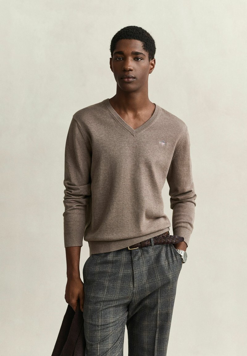 Jeune homme à la peau foncée porte un pull taupe à col en V et un pantalon à carreaux gris, tenant un manteau sombre dans une main, debout devant un fond neutre.