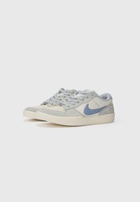 Šviesiai pilki sportbačiai, pagaminti iš zomšo ir audinio, su mėlynu Nike ženkliuku, standartine raištelių sistema ir guminėmis neslidiomis padangomis.