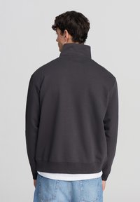 Felpa grigio carbone con colletto alto, maniche lunghe e vestibilità rilassata. Presenta polsini e orlo a coste. Indossata sopra una camicia bianca.