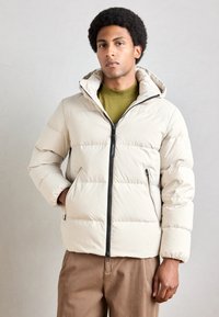 Puffer jacket in light beige with horizontal quilted design, zipper closure, hood, and side pockets. Worn over a green top and brown trousers.