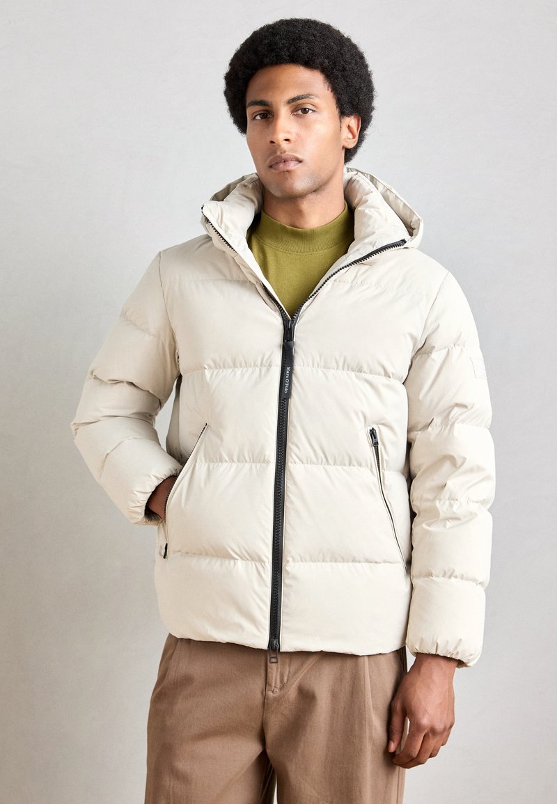 Puffer jacket in light beige with horizontal quilted design, zipper closure, hood, and side pockets. Worn over a green top and brown trousers.
