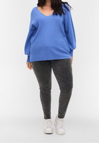 Blauer Strickpullover mit V-Ausschnitt, oversized Passform und Ballonärmeln, kombiniert mit grauen Skinny Jeans und weißen Sneakers.