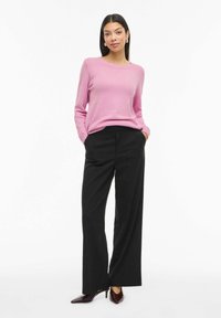 Gaiši rozā džemperis ar ribotu aproču apdari, kombinēts ar augsta vidukļa melnām plaša stila biksēm un sarkangaiļa toņa smailām kurpēm. Gluda tekstūra, ikdienas piegrieztne.