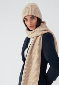 Juego de gorro y bufanda de punto beige, con textura acanalada. El gorro tiene un ajuste ceñido, mientras que la bufanda es larga y se encuentra drapeada sobre una chaqueta oscura.