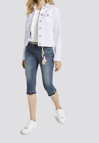 Chaqueta de mezclilla blanca con botones de presión, combinada con jeans de mezclilla azul recortados con dobladillos deshilachados y un cinturón de colores tejidos. Zapatillas blancas.