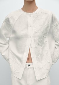 Donna che indossa un cardigan bianco a trama con bottoni, con ricami floreali, parzialmente aperto, abbinato a pantaloni bianchi a vita alta, su uno sfondo neutro.