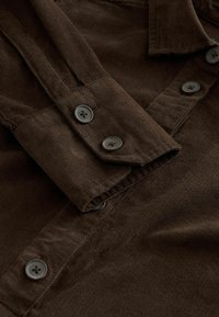 Kohverpuuvillane pruun särk, millel on klassikaline krae, nööbid mansettides ja tekstureeritud kangas. Märkimisväärsed detailid on ümmargused tumedad nööbid ja ribakujundus.