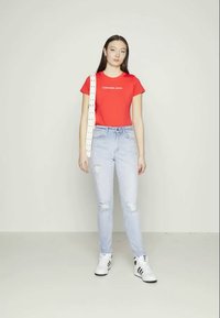 Camiseta curta vermelha com o logótipo "Calvin Klein Jeans", jeans claros desgastados e sapatilhas brancas com riscas pretas. Bolsa de ombro bege.