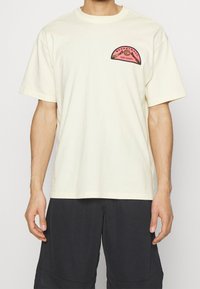 Krémové tričko s krátkým rukávem a kulatým výstřihem. Na hrudi má grafiku "Basketball Nike Hoops" v růžové a černé barvě.