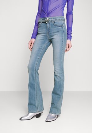 Person iført lyseblå vidde jeans og sølvfarvede spidse støvler, kombineret med en gennemsigtig lilla langærmet top, stående mod en ensfarvet baggrund.