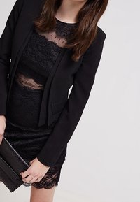 Černý blazer z texturovaného materiálu s přiléhavým střihem, kombinovaný s černými krajkovými šaty s průhledným efektem, které mají zubatý lem a odpovídající kabelku.