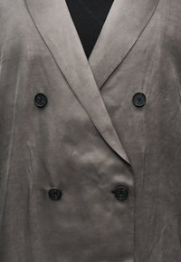 Κοντινό πλάνο γκρι διπλού breasted παλτού με μαύρα κουμπιά πάνω από μαύρο τοπ, που δείχνει γιακάδες και λεπτομέρειες κουμπιών.