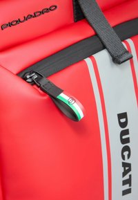 Mochila impermeable roja con herrajes negros, con una cremallera prominente y detalle de la bandera italiana en el tirador de la cremallera. Logotipo "DUCATI" mostrado.