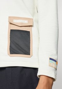 Top blanco de manga larga con relieve, con un bolsillo texturizado beige y un inserto de malla negra. Logotipo de parche en la manga con un diseño de montaña y sol.