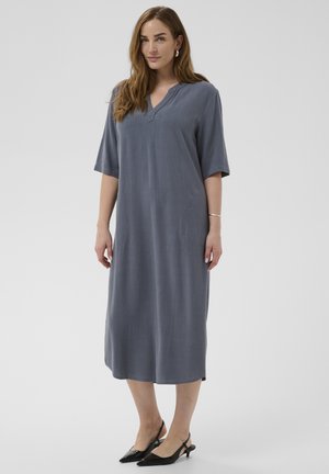 Woman wearing a loose, knee-length slate blue dress with short sleeves and black low-heeled slingback shoes, standing against a plain background.