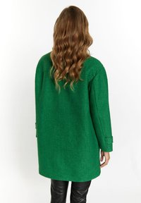 Cappotto verde testurizzato con una vestibilità rilassata, maniche lunghe e taglio dritto. Presenta un colletto arrotondato e tasche a toppa. Indossato con pantaloni neri.