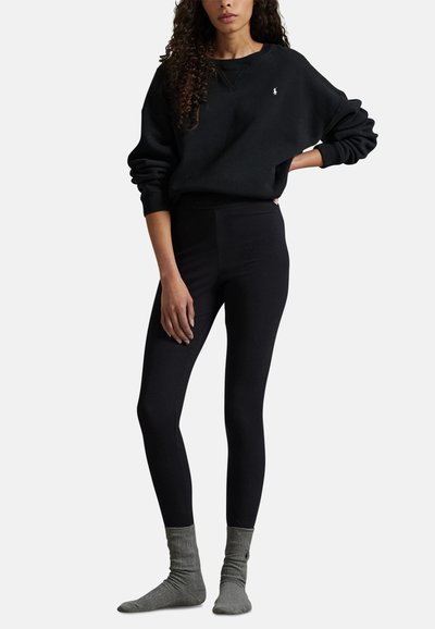 Černý svetr s malým logem na hrudi, v kombinaci s černými legínami a šedými žebrovanými ponožkami. Měkká tkanina, volný střih a jednobarevné provedení.