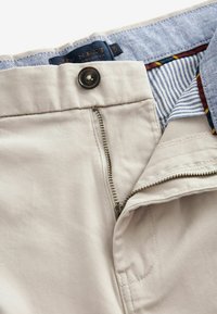 Pantalon beige à coupe droite avec braguette zippée, fermeture par bouton et ceinture intérieure rayée bleu et blanc. Tissu lisse avec des détails subtils de texture.