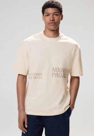 Krämfärgad bomulls-t-shirt med korta ärmar, med grafisk text i beige: "NOUVEAU PROJECT" och "DE CRÉMIEUX PARIS 75112."