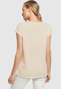 Beige kortærmet t-shirt lavet af blødt stof, med en let buet kant og oprullede ærmer, båret sammen med hvide bukser.