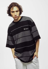Oversized zwart en grijs gestreepte t-shirt met korte mouwen, met een logo in een contrasterend lettertype op de linkerborst. Te combineren met grijze joggingbroek.