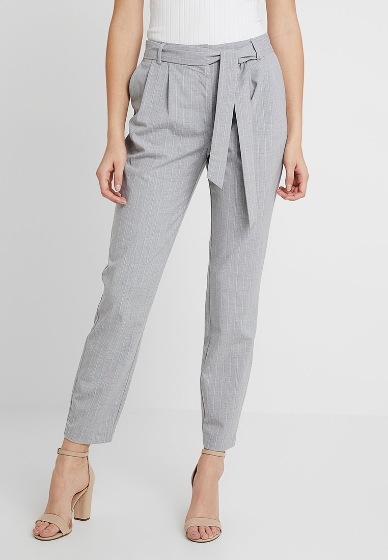 Světle šedé kalhoty na míru s vertikálními proužky, plissé přední částí a opaskem. Vyrobené z hladké tkaniny s tapered nohavicemi.
