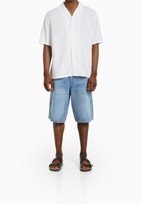 Man som bär en vit kortärmad skjorta med knappar, ljusblå denimshorts och svarta sandaler med spänne, som står framför en vit bakgrund.