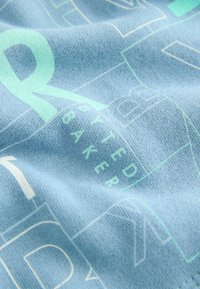 Svetlomodrá textília s jemnou štruktúrou, na ktorej sú po celom povrchu zobrazené veľké, výrazné mintovozelené a biele grafické písmená a číslice.