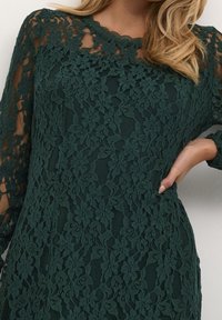 Femme en robe en dentelle fleurie vert foncé à manches longues, main posée sur la hanche, cheveux blonds ondulés tombant sur l'épaule droite.