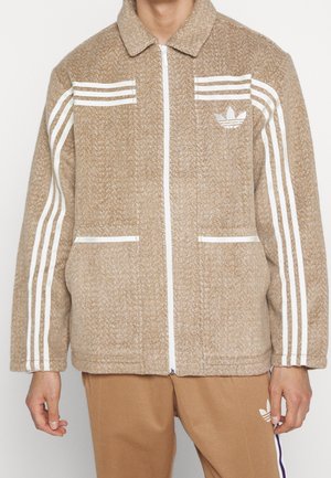 Mann trägt eine hellbraune Jacke mit Reißverschluss, weiße Adidas-Streifen an den Ärmeln und ein Logo auf der Brust, kombiniert mit passenden braunen Hosen.