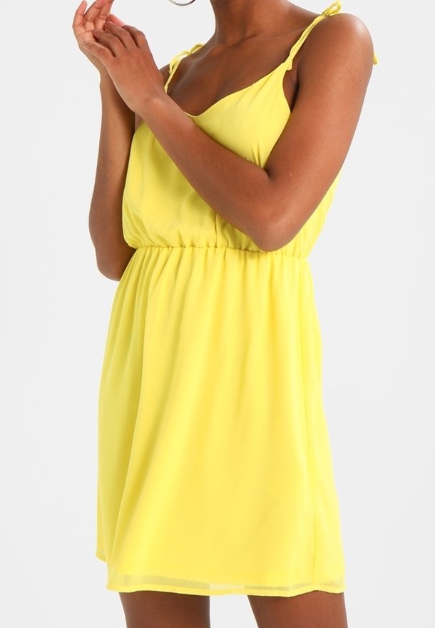 Femme portant une robe jaune sans manches avec des bretelles à nouer, taille cintrée, debout les mains jointes près du visage sur un fond uni.
