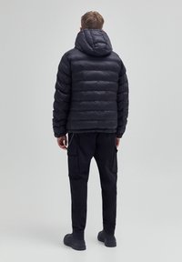 Hombre de pie de espaldas, vistiendo una chaqueta acolchada negra con capucha, pantalones cargo negros y zapatos negros, sobre un fondo gris liso.