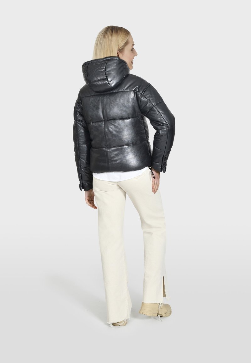 Schwarze Steppjacke mit glänzendem Finish, Kapuze und Reißverschlussdetails, kombiniert mit off-white weit geschnittenen Hosen und beigen Sneakers.
