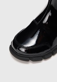 Γυαλιστερό μαύρο δερμάτινο μποτάκι με ανθεκτική καουτσούκ σόλα και λεπτομερή ραφή σε λευκό φόντο.