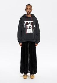 Hoodie oversize nero con grafica logo argentata e lacci rossi, abbinato a una lunga gonna nera plissettata e sneaker beige.