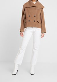 Brun ylleblandning dubbelknäppt jacka med stor krage, fickor med lappar och svarta knappar, tillsammans med vita utsvängda byxor och vita klackstövlar.