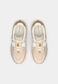 Baskets beige clair avec une tige en maille texturée, des accents blancs et une bande beige sur le talon. Elles présentent un design à lacets plats et un logo sur la languette.