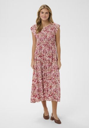 Mujer de pie que lleva un vestido midi sin mangas con estampado floral en tonos rosas y rojos, con pliegues, y zapatos marrones de tacón bloque y tejido.