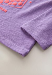 Lilla lühikese varrukaga särk pehme tekstuuriga, millel on punane graafikadisain ja kontrastsed õmblused ääres ja varrukate servades.