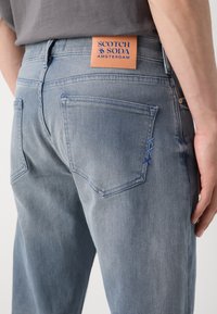Jeans en denim bleu clair avec un détail de couture bleue sur la poche arrière et une étiquette en cuir beige portant la mention "Scotch & Soda Amsterdam."