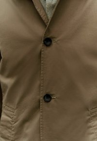 Giacca button-up marrone con una texture liscia, caratterizzata da due bottoni neri e una tasca laterale. Sotto, è visibile un maglione di lana grigio chiaro.