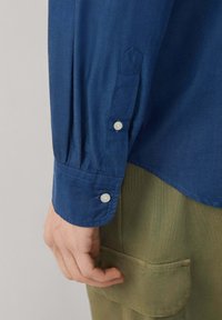 Camisa azul de botones con una textura suave, que presenta un puño puntiagudo y botones blancos. Combinada con pantalones verde oliva, que destacan por un detalle de bolsillo.